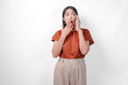 Shocked Asian woman in a brown shirt gesturing whisper to someone with her hand while covering her mouth, isolated by white background.の写真素材