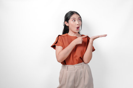 Pretty young Asian woman in a brown shirt and taupe pants is pointing to the copy space beside her with shocked expression, isolated by white background.の写真素材