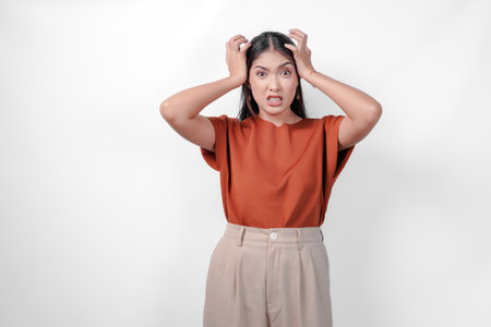 Stressed young Asian woman in a brown shirt and taupe pants is holding her head in her hands suffering from headache.の写真素材