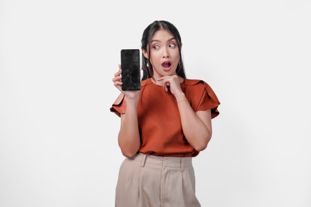 Surprised young Asian woman in brown shirt showing the copy space on phone screen, isolated white background.の写真素材