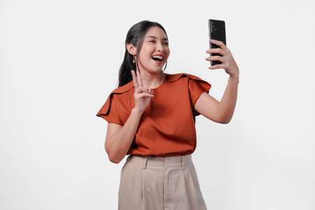 Excited young Asian woman in a brown shirt is taking a selfie with her cell phone with peace finger gesture over isolated white background.の写真素材