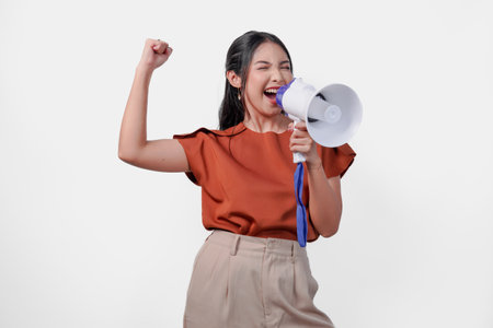 Happy successful Asian woman wearing a brown shirt raising clenched fist while shouting and screaming loud using megaphone speaker. Communication concept.の写真素材