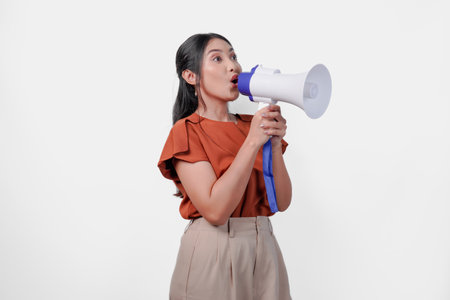 Lively young Asian woman shouting at megaphone. Concept of excitement and energy, as if the woman is performing or giving a speech.の写真素材