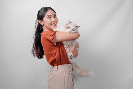 Pretty young Asian woman in a brown shirt is holding a white ragdoll cat and raising it high over isolated white background.の写真素材