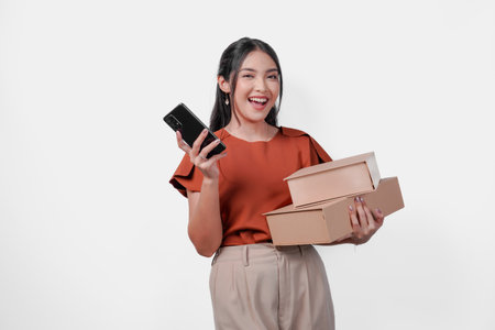 Excited Asian woman is holding a cell phone and boxes while smiling happily. Concept of excitement and joy, possibly related to receiving a package or communicating with someone.の写真素材