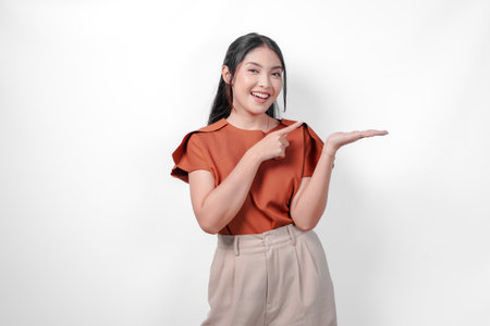 Smiling Asian woman in a brown shirt and taupe pants is pointing to the copy space beside her, isolated by white background.の写真素材