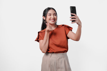 Lovely young Asian woman in a brown shirt is doing video call and showing finger heart gesture to the camera, isolated by white background.の写真素材
