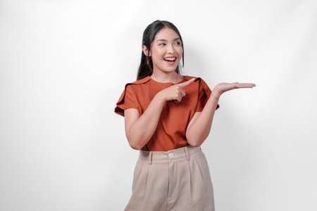 Smiling Asian woman in a brown shirt and taupe pants is pointing to the copy space beside her, isolated by white background.の写真素材