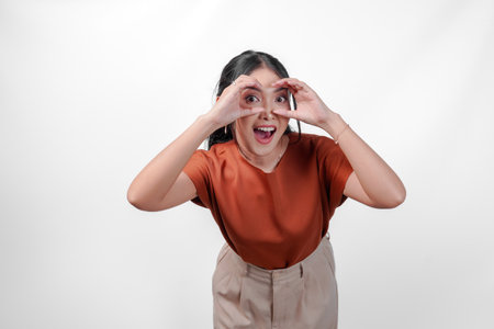 Happy young Asian woman wearing a brown shirt making binocular gesture with her hands over isolated white background.の写真素材