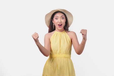 Joyful young Asian woman in a yellow dress holding her hands up in the air with a happy successful expression. She is wearing a straw hat and earrings.の写真素材