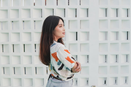 Chic Asian woman wearing a colorful shirt stands in front of a white wall with crossed arms while...