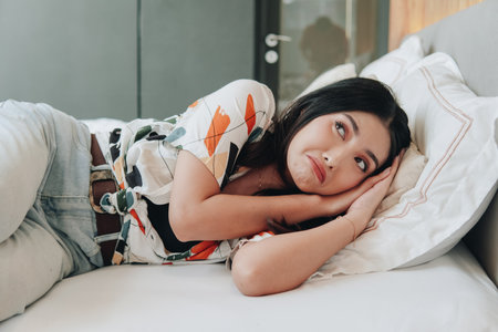 Anxious young Asian woman wearing bright denim and patterned shirt is laying on a bed feeling uneasy...