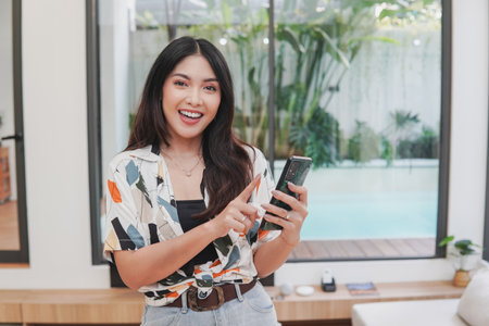 Joyful young Asian woman is smiling and holding smartphone, posing in a room with swimming pool outside.の写真素材