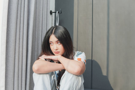Serious Asian woman is sitting on the floor with her hands on her knees, looking to the side while in a deep thought.の写真素材