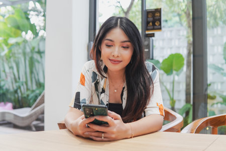 Young Asian woman is sitting at a dinner table while holding smartphone and looking with a smile.の写真素材