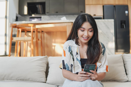 Excited young Asian woman is sitting on a couch and looking at her cell phone with villa kitchen island in the background.の写真素材
