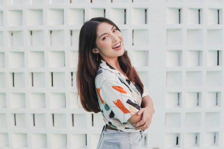 Cheerful young Asian woman is smiling and wearing a white shirt with a black and orange pattern. She is standing in front of a white wall.の写真素材
