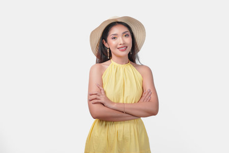 Confident young Asian woman in a yellow dress is wearing a straw hat standing with her arms crossed and looking at the camera, isolated by white background.の写真素材