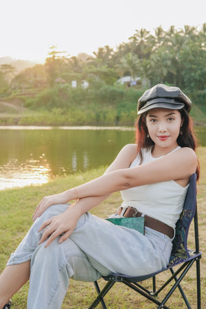 Smiling woman is sitting in a lawn chair by a lake looking at the camera. She is wearing a black hat...
