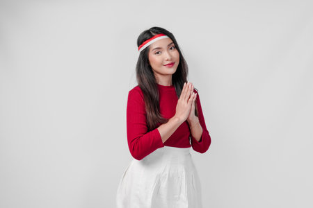 Calm young Asian woman wearing a red shirt and white skirt is smiling and doing traditional greeting gesture. Indonesia Independence Day concept.の写真素材