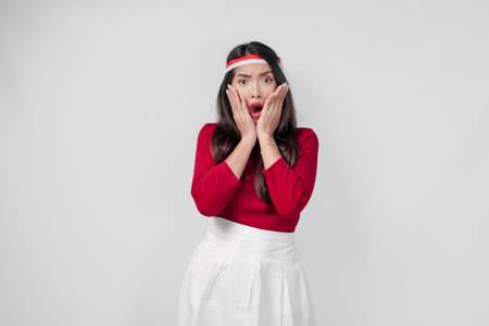 Young Asian woman in a red shirt and white skirt is looking surprised while covering her mouth.の写真素材