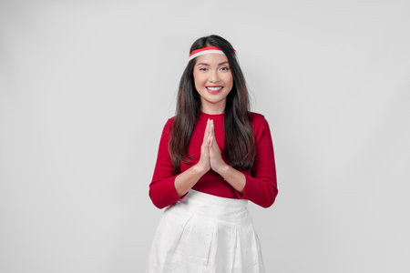 Calm young Asian woman wearing a red shirt and white skirt is smiling and doing traditional greeting gesture. Indonesia Independence Day concept.の写真素材