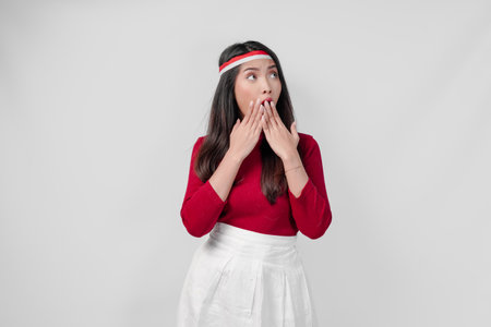 Young Asian woman in a red shirt and white skirt is looking surprised while covering her mouth.の写真素材