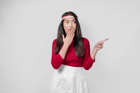 Surprised young woman in a red shirt and white pants is pointing to the copy space beside her, isolated white background.の写真素材