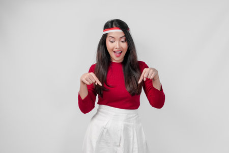 Joyful young woman in a red shirt and white pants is pointing to the copy space downward, isolated white background.の写真素材