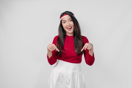 Joyful young woman in a red shirt and white pants is pointing to the copy space downward, isolated...