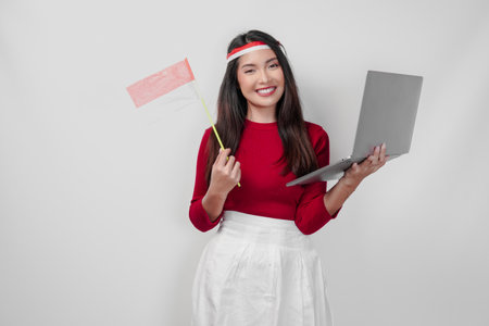Joyful young Asian woman is holding a laptop and a mini country flag. Indonesia Independence Day concept.の写真素材