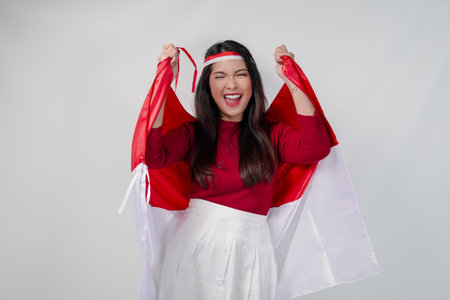 Cheerful young Asian woman is holding Indonesian flag at her back and raising fists in the air with happy successful expression. Indonesian Independence Day concept.の写真素材