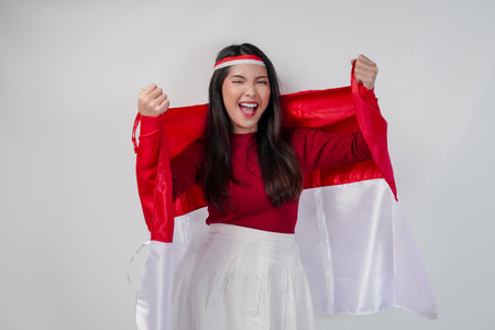 Cheerful young Asian woman is holding Indonesian flag at her back and raising fists in the air with happy successful expression. Indonesian Independence Day concept.の写真素材