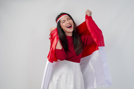 Cheerful young Asian woman is holding Indonesian flag at her back and raising fists in the air with happy successful expression. Indonesian Independence Day concept.の写真素材