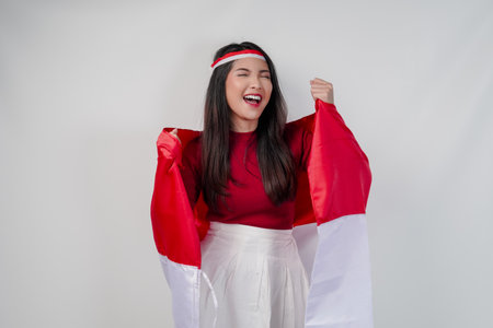 Cheerful young Asian woman is holding Indonesian flag at her back and raising fists in the air with happy successful expression. Indonesian Independence Day concept.の写真素材