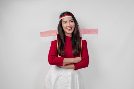 A happy young Asian woman is holding two mini Indonesian flags and smiling with crossed arms gesture of confidence. Indonesia Independence Day concept.の写真素材