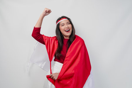 Happy young Asian woman with Indonesia flag draped over her shoulders and raising fist hand gesture. Indonesia Independence Day concept.の写真素材
