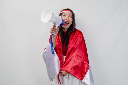 Energetic Asian woman with country flag draped over her shoulder while shouting at a megaphone. Indonesia Independence Day concept.の写真素材