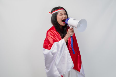 Energetic Asian woman with country flag draped over her shoulder while shouting at a megaphone.の写真素材
