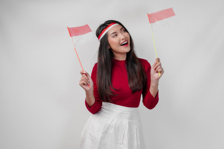 Joyful young Asian woman is holding two flags and smiling, isolated by white background. Indonesia Independence Day concept.の写真素材