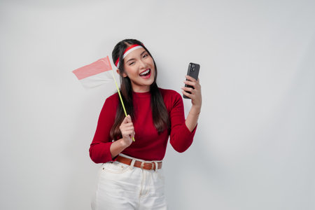 Excited young Asian woman holding Indonesian mini flag smiling while using her cell phone. Indonesia Independence Day concept.の写真素材