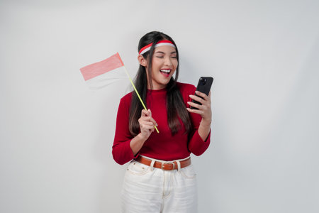 Excited young Asian woman holding Indonesian mini flag smiling while using her cell phone. Indonesia Independence Day concept.の写真素材