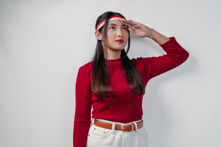 Beautiful young Asian woman in a red shirt and white pants doing salute pose to country flag. Indonesia Independence Day concept.の写真素材