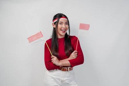 A happy young Asian woman is holding two mini Indonesian flags and smiling with crossed arms gesture of confidence. Indonesia Independence Day concept.の写真素材