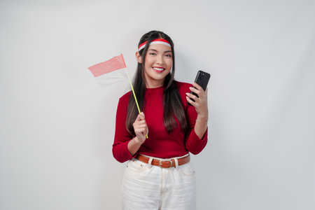 Excited young Asian woman holding Indonesian mini flag smiling while using her cell phone. Indonesia...