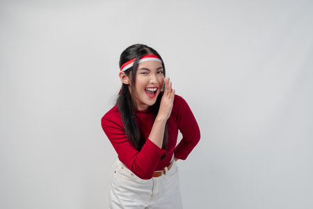 Cute young Asian woman in a red shirt and white pants is making whisper gesture with hand, isolated by white background. Indonesia Independence Day concept.の写真素材