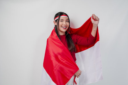 Confident Asian woman in a red and white outfit is covering herself with Indonesian flag and raising a fist. Indonesia Independence Day concept.の写真素材
