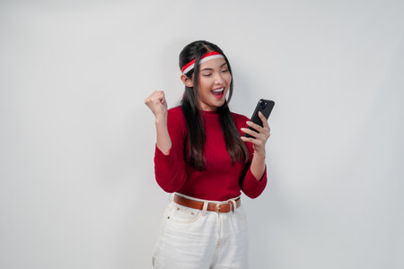 Excited Asian woman in a red shirt holding a cell phone and while raising fist to celebrate victory or a happy moment.の写真素材