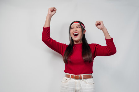 Cheerful young Asian woman in a red shirt and white pants is smiling and raising her hand in the air.の写真素材