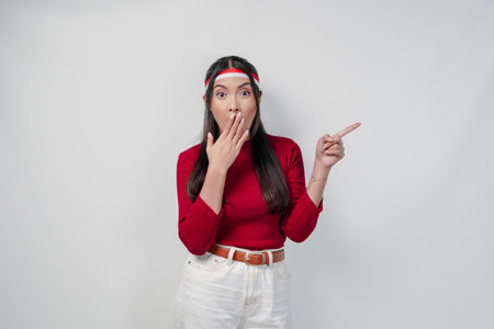 Surprised young woman in a red shirt and white pants is pointing to the copy space beside her, isolated white background. Indonesia Independence Day concept.の写真素材
